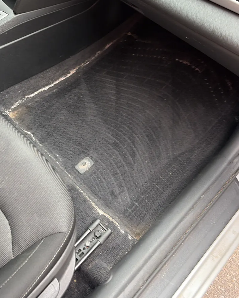 Passenger footwell of a sedan before cleaning, floor mat greyed with dried road salt and ground-in dirt along the edges